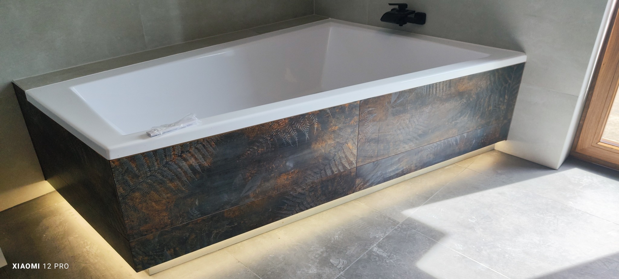 Wanna obudowana płytkami z motywem paproci, podświetlenie LED u dołu, szara płytka podłogowa i ścienna, czarna bateria wannowa.