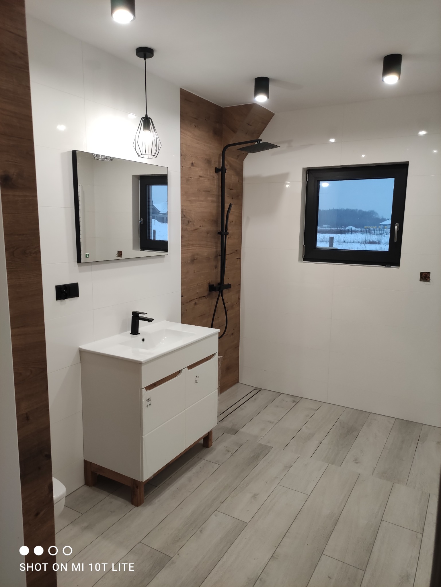 Nowoczesna łazienka z białymi płytkami, drewnopodobną ścianą i podłogą z płytek imitujących drewno, czarną armaturą, wiszącą lampą i oknem z widokiem na zimowy krajobraz.