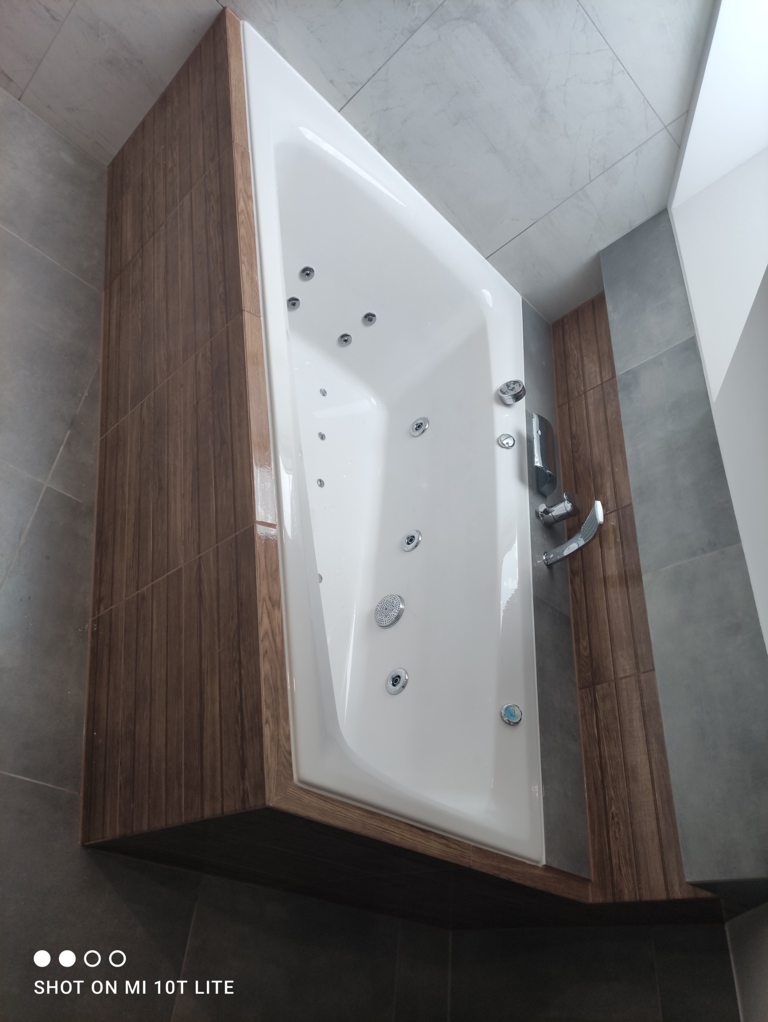 Biała wanna z hydromasażem, obudowana panelami imitującymi drewno, z chromowaną armaturą, na tle szarych płytek w łazience.