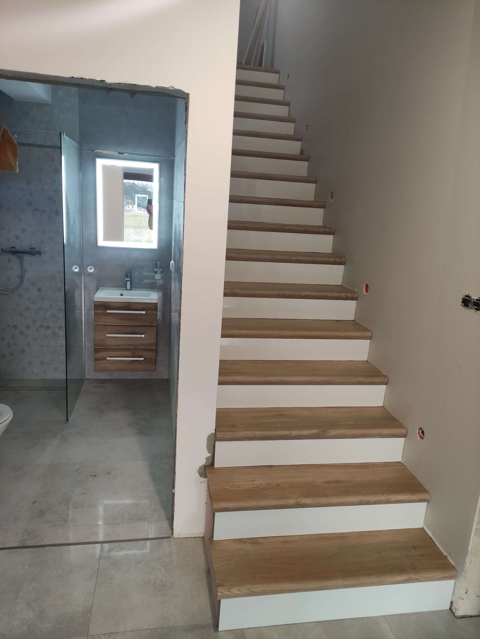 Wnętrze w trakcie prac: Schody z drewnianymi stopniami i białymi podstopnicami, obok wejście do łazienki z szarą glazurą, umywalką z szafką z drewnopodobnym fornirem i lustrem z podświetleniem LED.