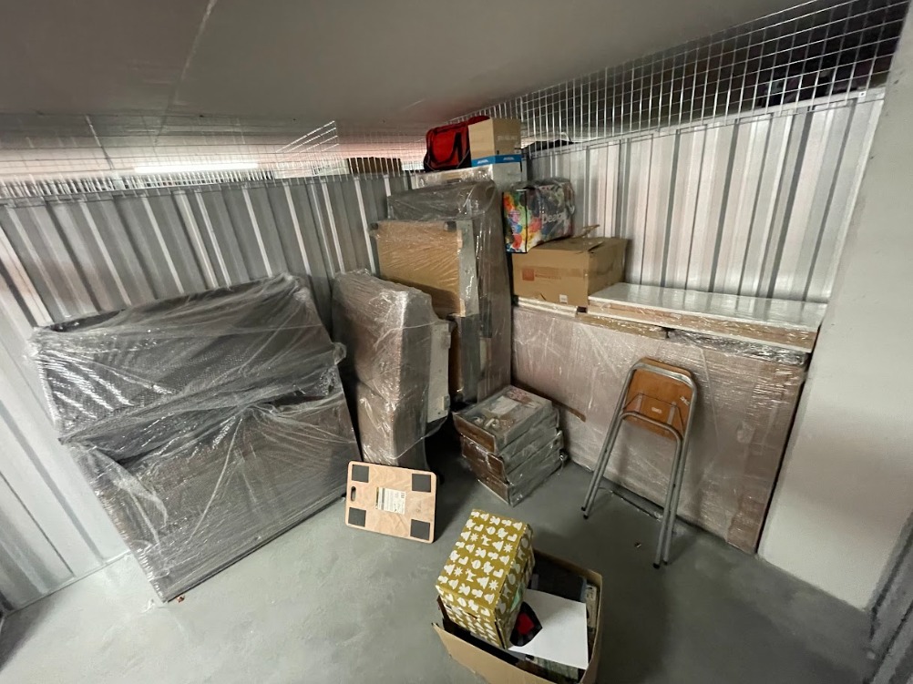 Zapakowane meble i kartony w magazynie tymczasowym z metalowymi ścianami. Krzesło i wózek transportowy widoczne. Oświetlenie sufitowe.