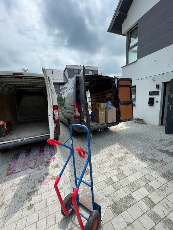 Dwa busy z otwartymi bagażnikami na posesji przed domem. W tle kartony, a na pierwszym planie wózek transportowy. Usługa przeprowadzkowa w trakcie realizacji.