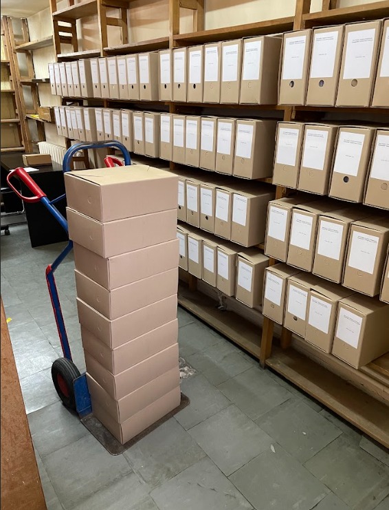 Wózek transportowy z wysokim stosem kartonów na tle regałów z segregatorami w archiwum. Przygotowanie do przeprowadzki biura lub opróżniania magazynu.