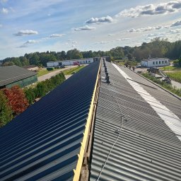 USŁUGI BUDOWLANE ARKADIUSZ PESTKA - Długi dach z blachy falistej, częściowo wymieniony, z widoczną drewnianą konstrukcją i nowym ociepleniem, na tle budynków gospodarczych i błękitnego nieba z chmurami.