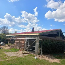 USŁUGI BUDOWLANE ARKADIUSZ PESTKA - Prace dekarskie w toku: dekarz na dachu z czerwonej dachówki mocuje elementy konstrukcyjne, widoczne rusztowania i przygotowane materiały budowlane na tle błękitnego nieba z chmurami.