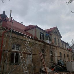 USŁUGI BUDOWLANE ARKADIUSZ PESTKA - Renowacja zabytkowego budynku z nową czerwoną dachówką, widoczne rusztowania, częściowo odnowiona elewacja i elementy dekoracyjne na dachu.