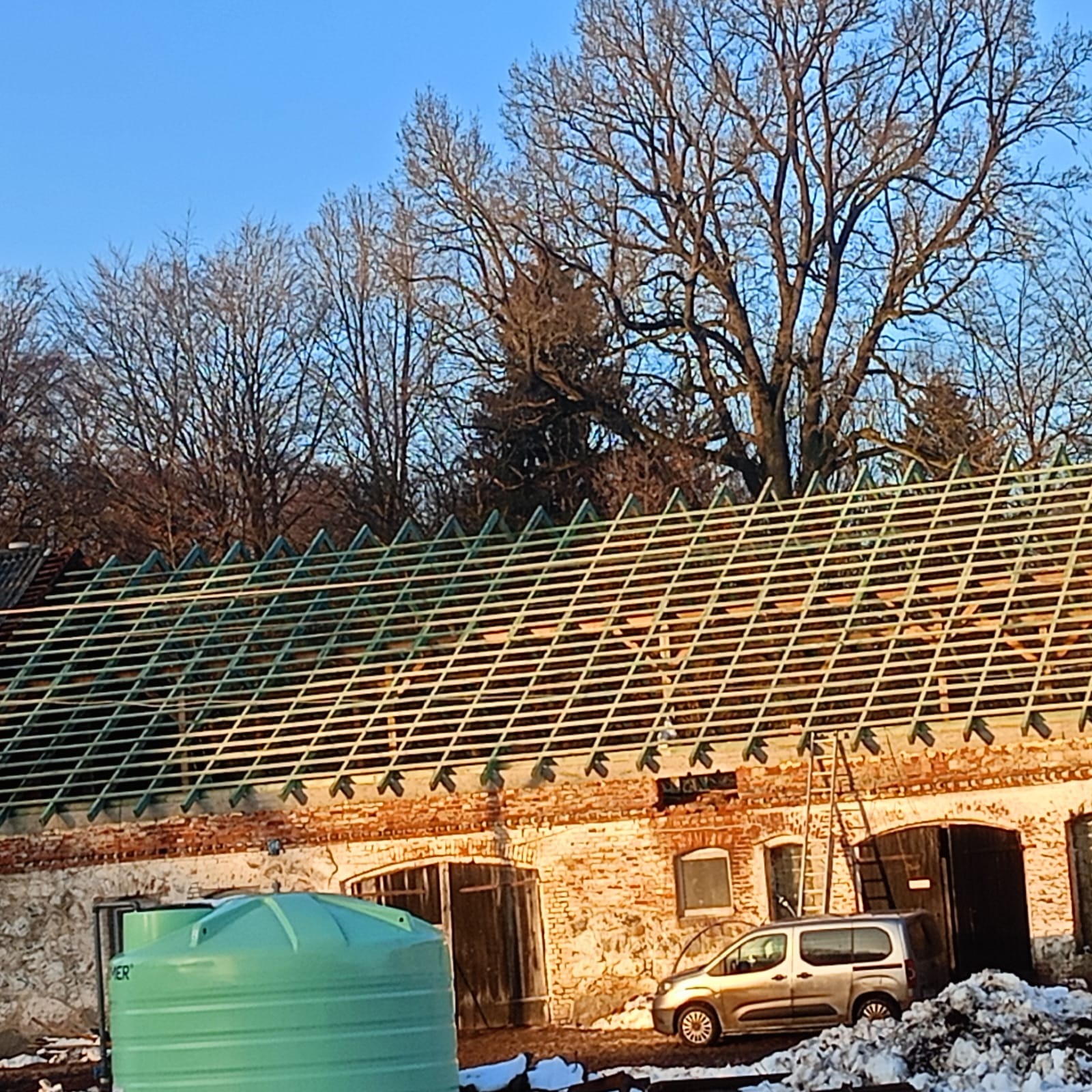 Konstrukcja więźby dachowej na ceglanym budynku gospodarczym, widoczna drabina oparta o ścianę, samochód dostawczy i zbiornik na wodę w zimowej scenerii.