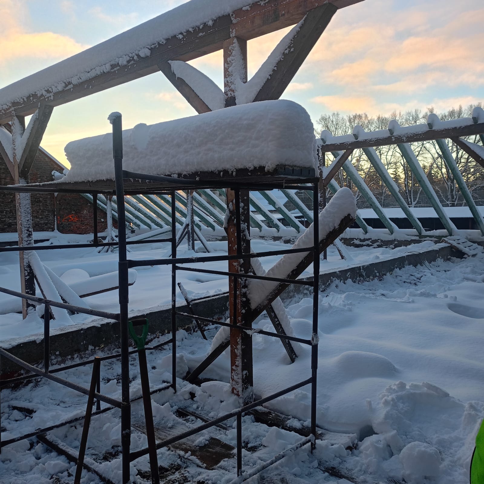 Śnieżna więźba dachowa w trakcie budowy, widok z rusztowania, drzewa w tle.