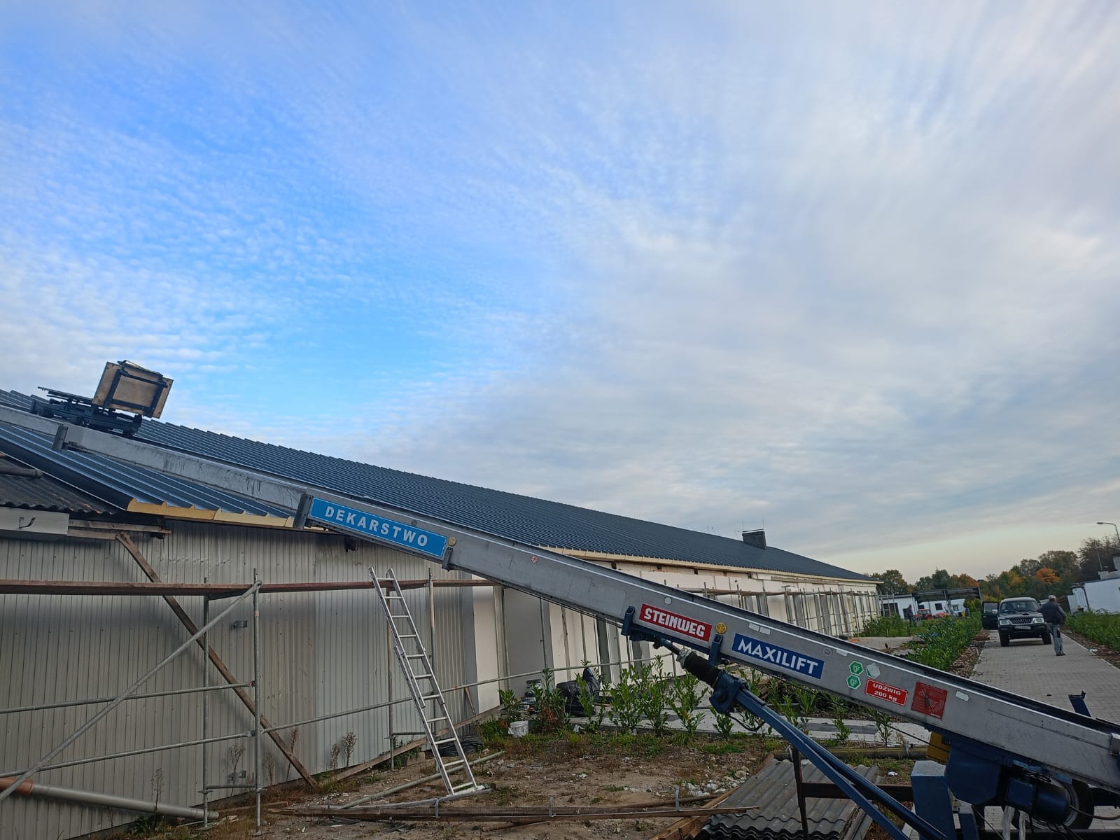 Prace dekarskie w toku: podnośnik Steinweg Maxilift ustawiony przy budynku z szarym dachem, widoczne rusztowania i drabina.