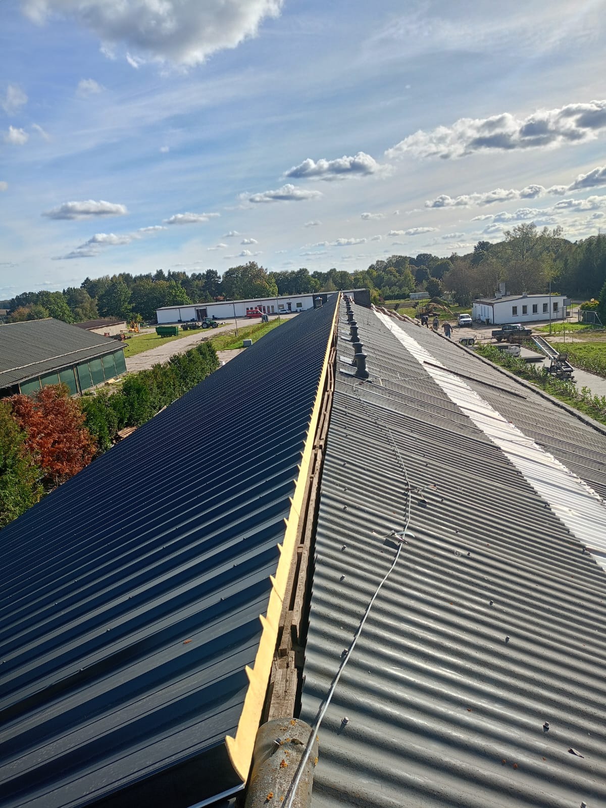 Długi dach z blachy falistej, częściowo wymieniony, z widoczną drewnianą konstrukcją i nowym ociepleniem, na tle budynków gospodarczych i błękitnego nieba z chmurami.