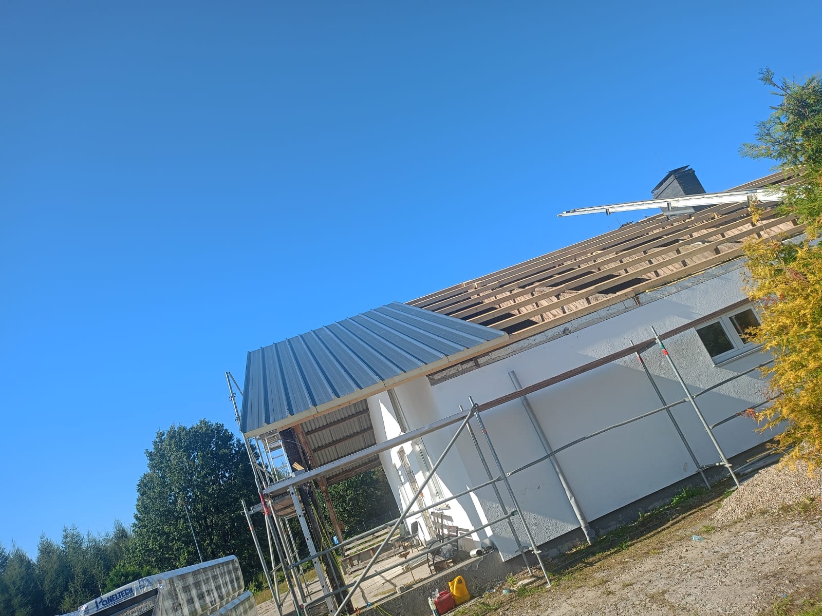 Dom w trakcie wymiany pokrycia dachowego, widoczne rusztowania, częściowo położona blacha trapezowa i więźba dachowa pod błękitnym niebem.
