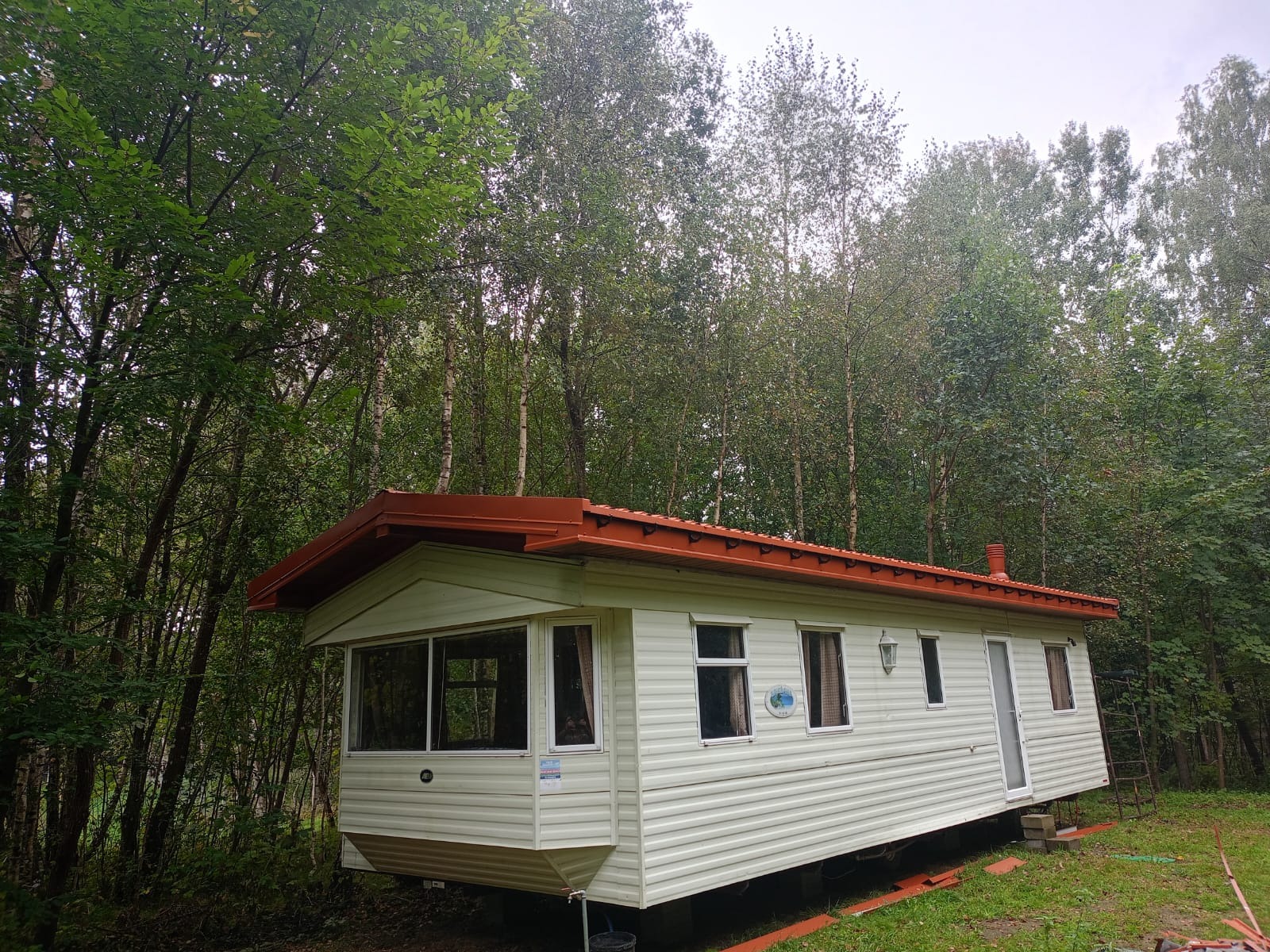 Biały, mobilny dom z pomarańczowym dachem, otoczony gęstym lasem.