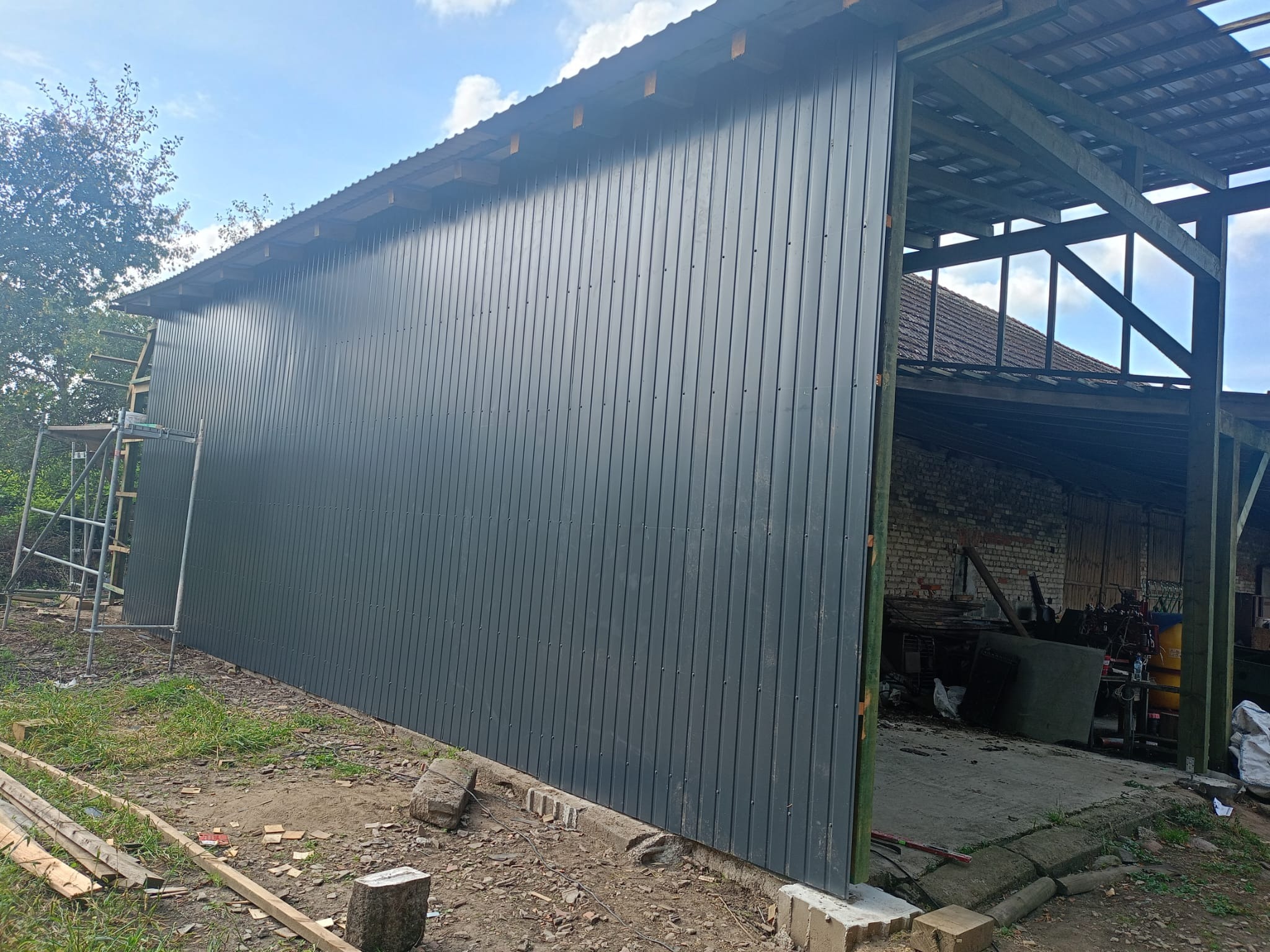 Nowa, szara blacha trapezowa na ścianie szopy, widoczna drabina i elementy konstrukcji dachu, w tle ceglany budynek gospodarczy.