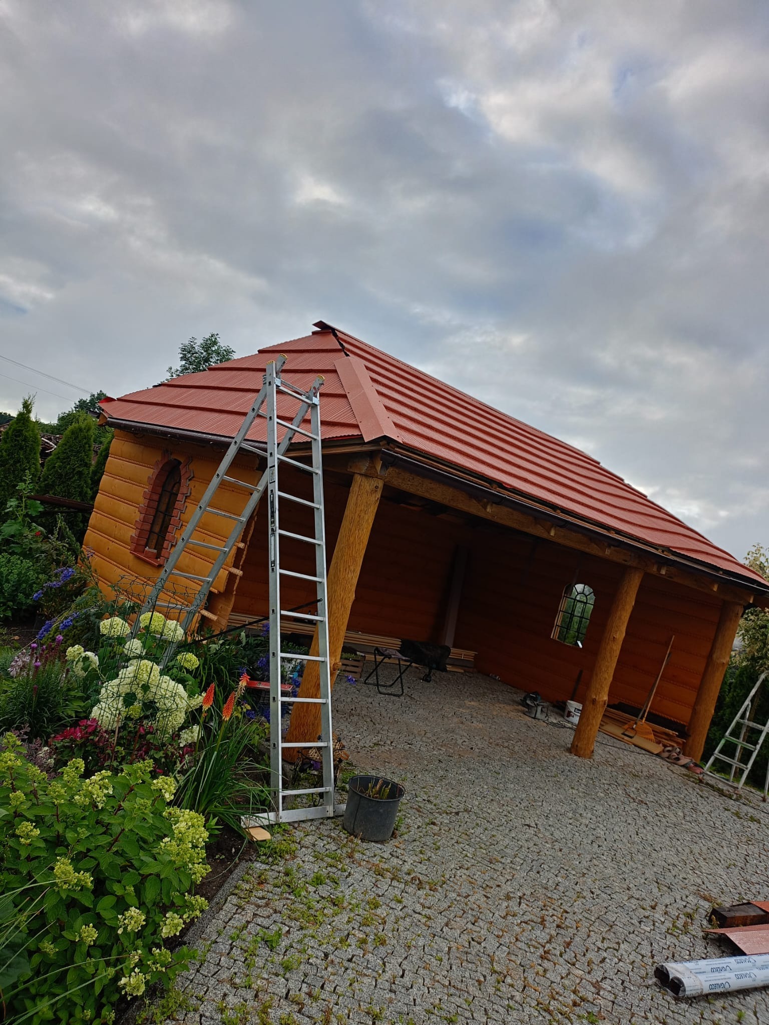 Drewniana altana z czerwoną dachówką w trakcie budowy, widoczna drabina oparta o ścianę, otoczenie to ogród z kwiatami i krzewami, na ziemi kostka brukowa.