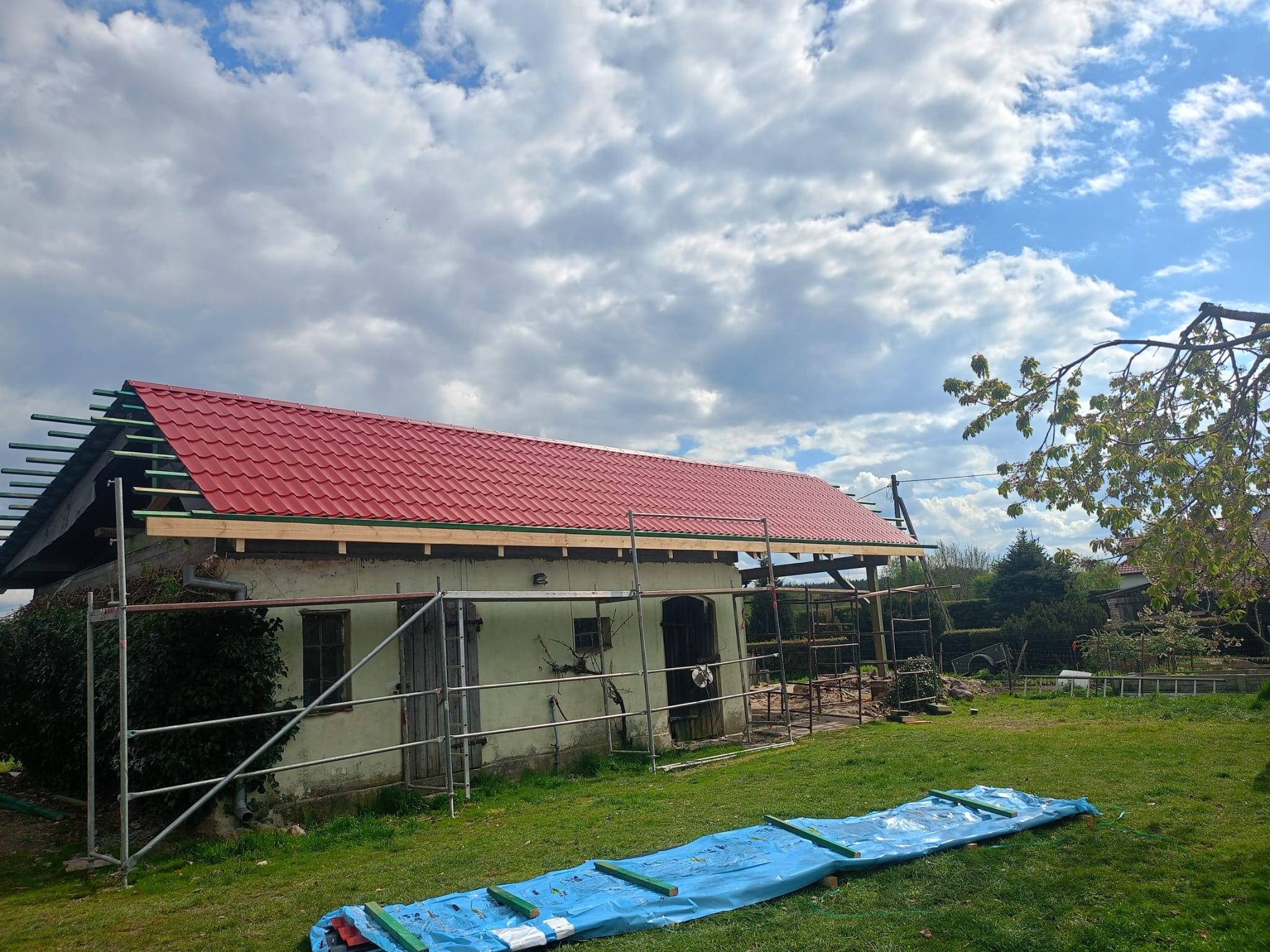 Budynek gospodarczy z częściowo wymienionym czerwonym dachem, widoczne rusztowania i przygotowane łaty pod dachówki oraz niebieska plandeka na trawie.
