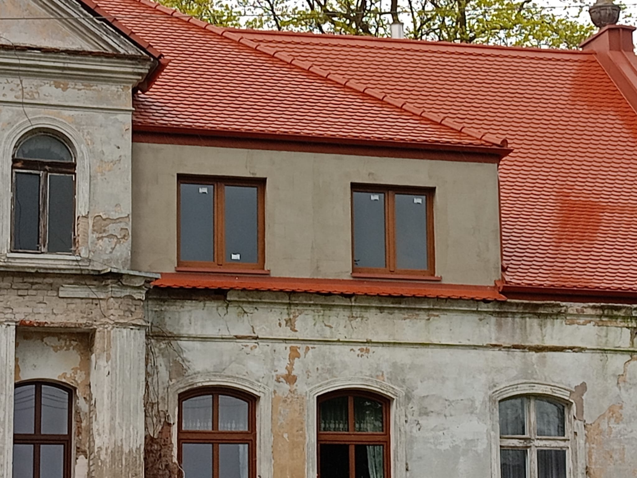Fragment elewacji budynku z nową, czerwoną dachówką i oknami, kontrastujący ze starą, łuszczącą się farbą na fasadzie.