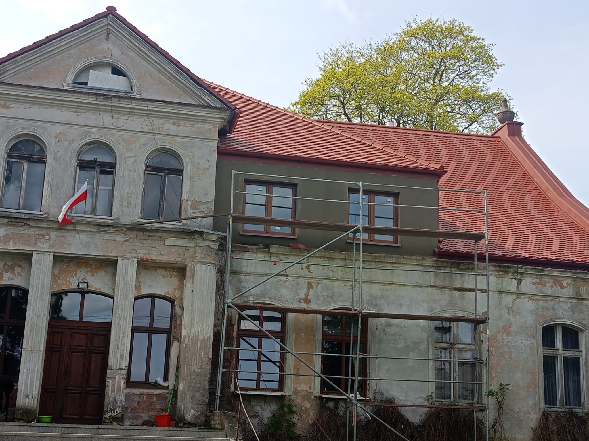 Renowacja zabytkowego dworku w Szczecinku: widoczne rusztowania, częściowo odnowiona elewacja, nowy dach z czerwonej dachówki, polska flaga w oknie, budynek w trakcie prac remontowych.