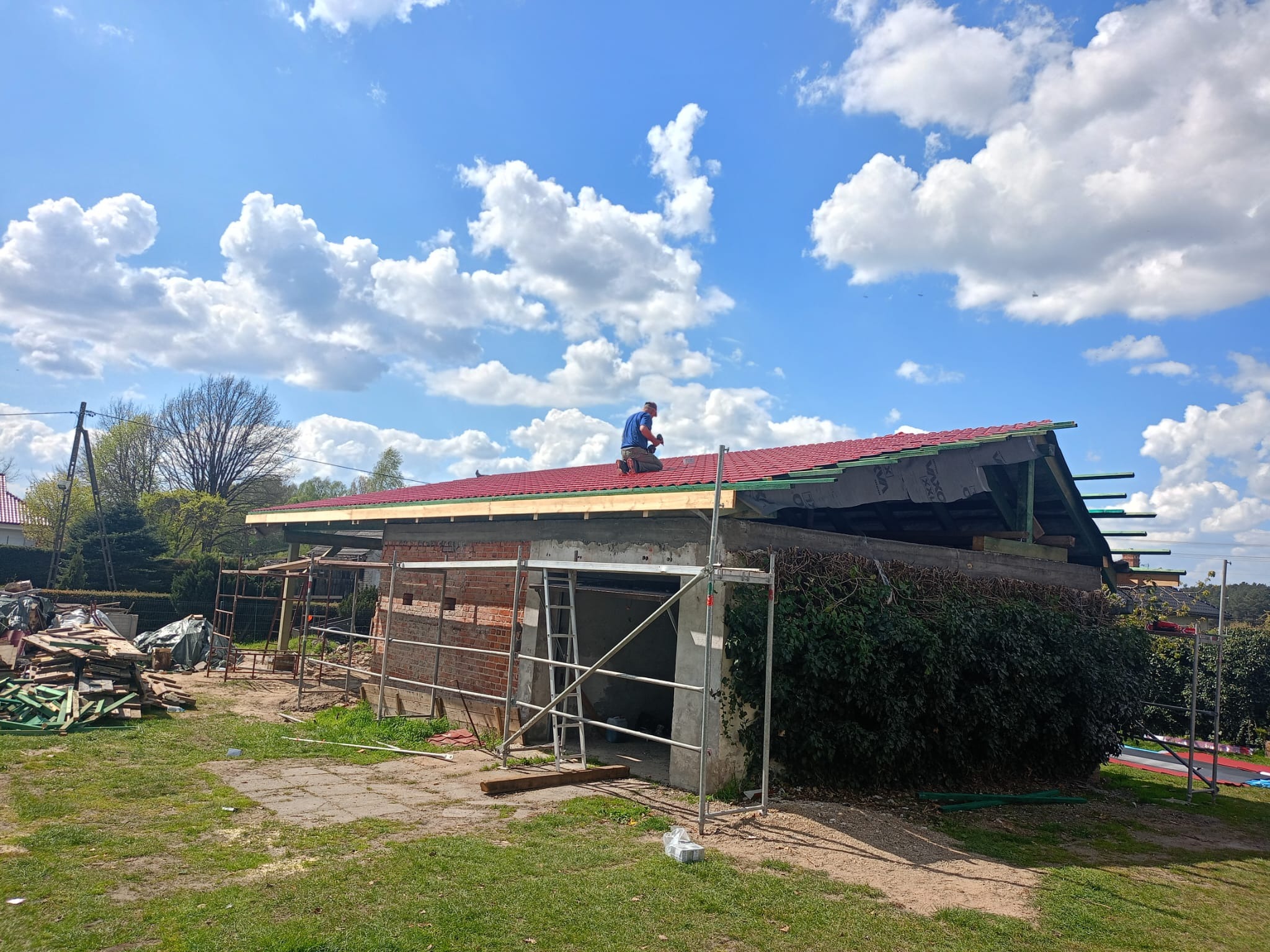Prace dekarskie w toku: dekarz na dachu z czerwonej dachówki mocuje elementy konstrukcyjne, widoczne rusztowania i przygotowane materiały budowlane na tle błękitnego nieba z chmurami.