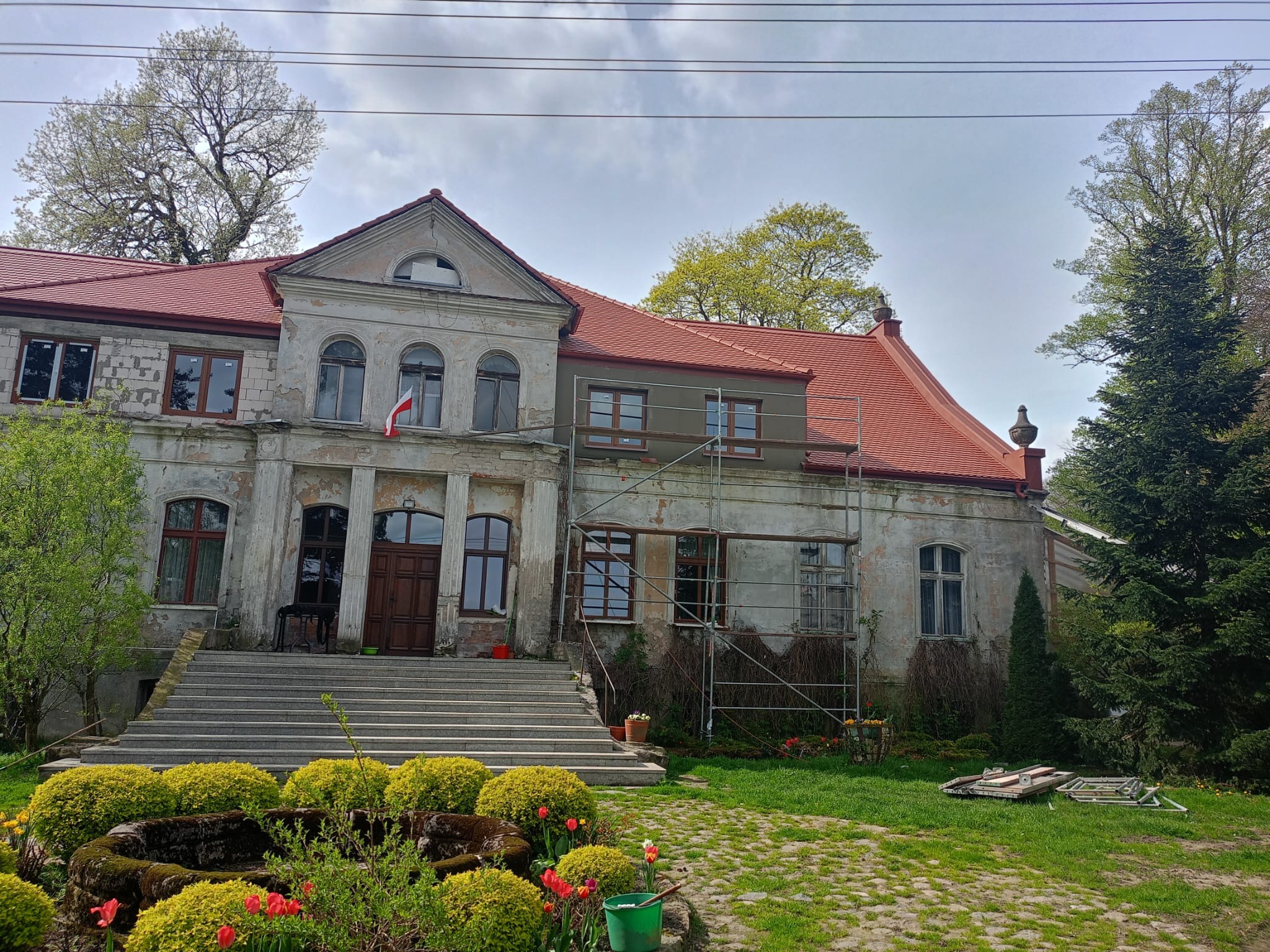 Renowacja zabytkowego dworku z nową czerwoną dachówką, widoczne rusztowania przy elewacji i flaga Polski w oknie.