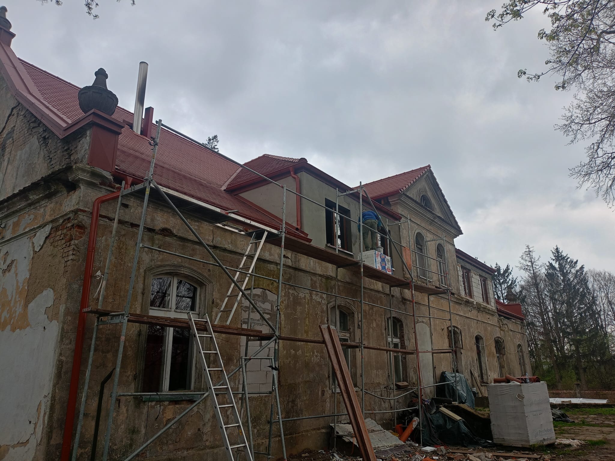 Renowacja zabytkowego budynku z nową czerwoną dachówką, widoczne rusztowania, częściowo odnowiona elewacja i elementy dekoracyjne na dachu.
