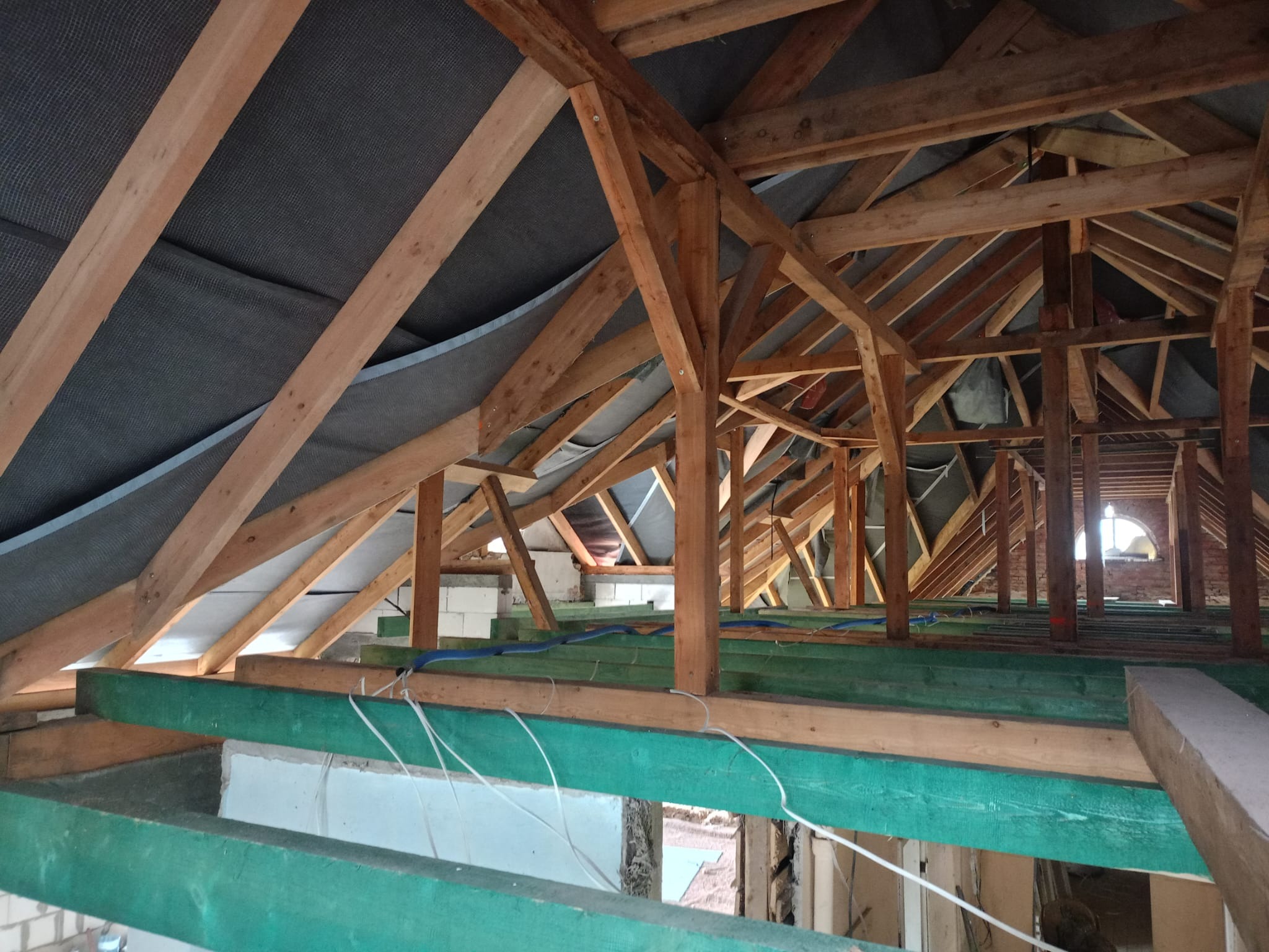 Surowa konstrukcja dachu z drewnianymi belkami, widoczna izolacja i fragment muru z oknem w tle, w trakcie budowy lub remontu.