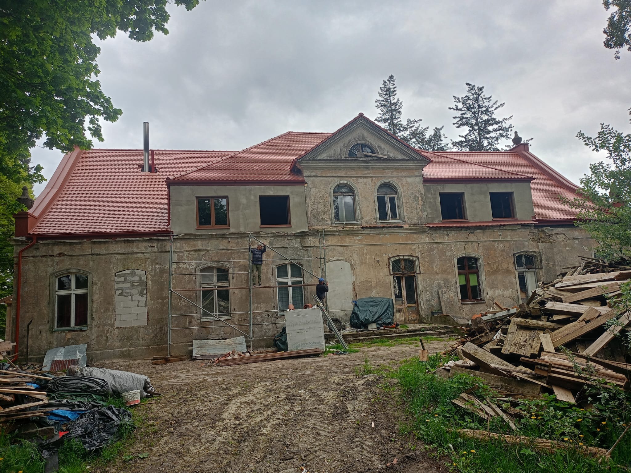 Renowacja starego pałacu: nowa, czerwona dachówka i pracownicy na rusztowaniu przy elewacji, obok sterta drewnianych odpadów.