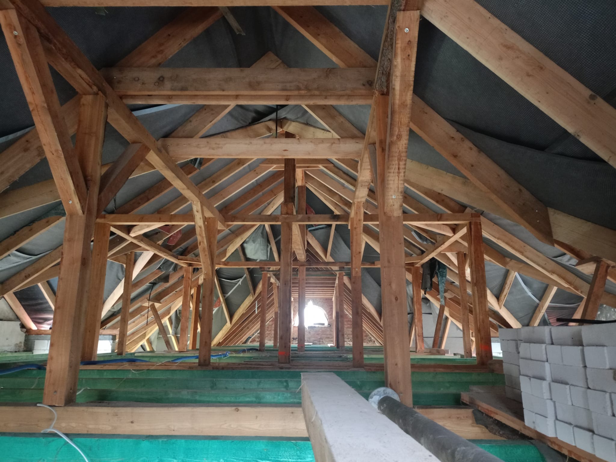 Surowa konstrukcja drewniana dachu od wewnątrz, widoczne elementy więźby, membrana dachowa oraz przygotowane materiały budowlane, w tle okno w murze.