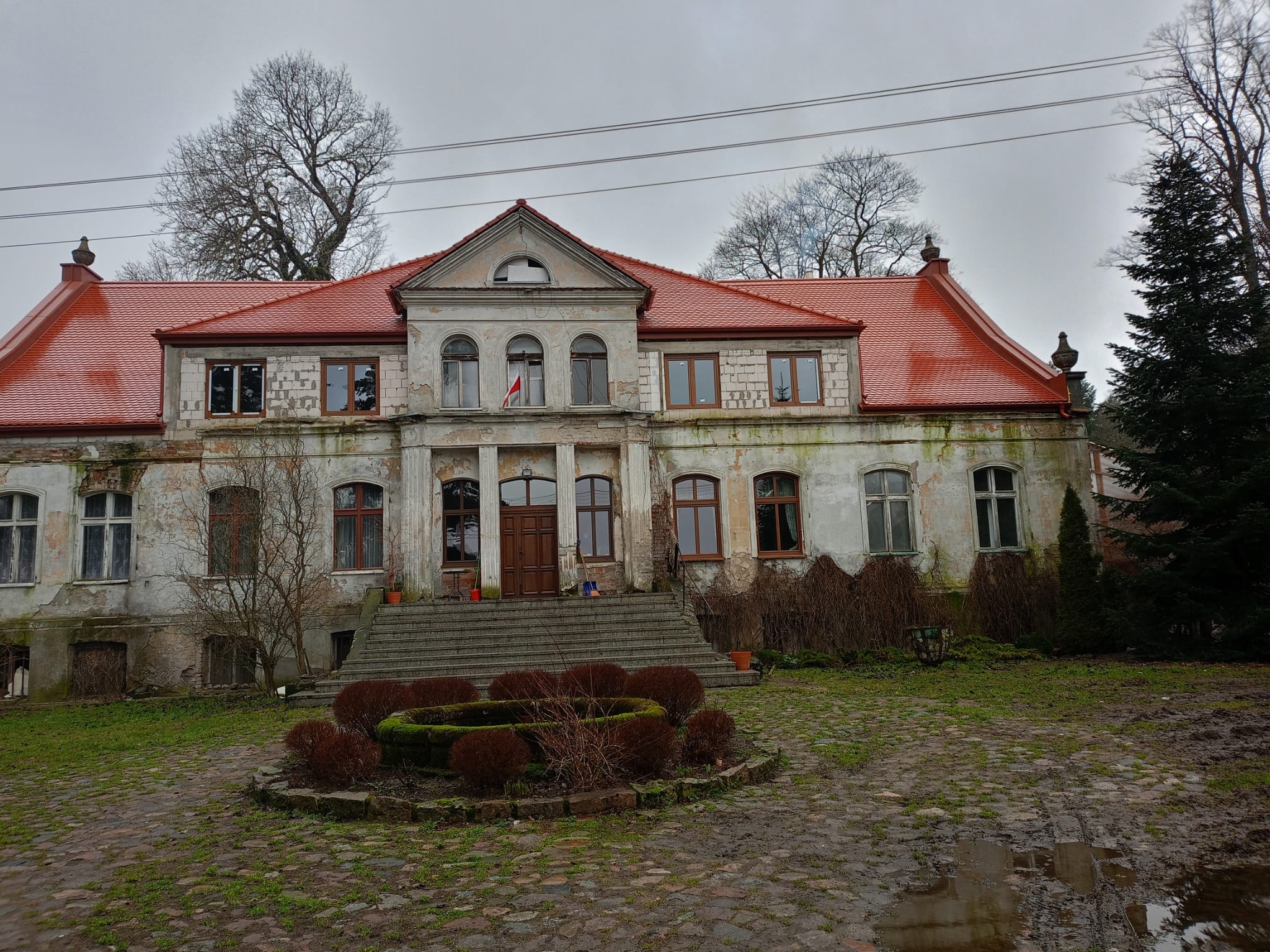Zrujnowany dwór z czerwoną dachówką, widoczne ślady zaniedbania i erozji na elewacji, na dachu miejscami brakuje dachówek, otoczenie zaniedbane z błotnistym podłożem.
