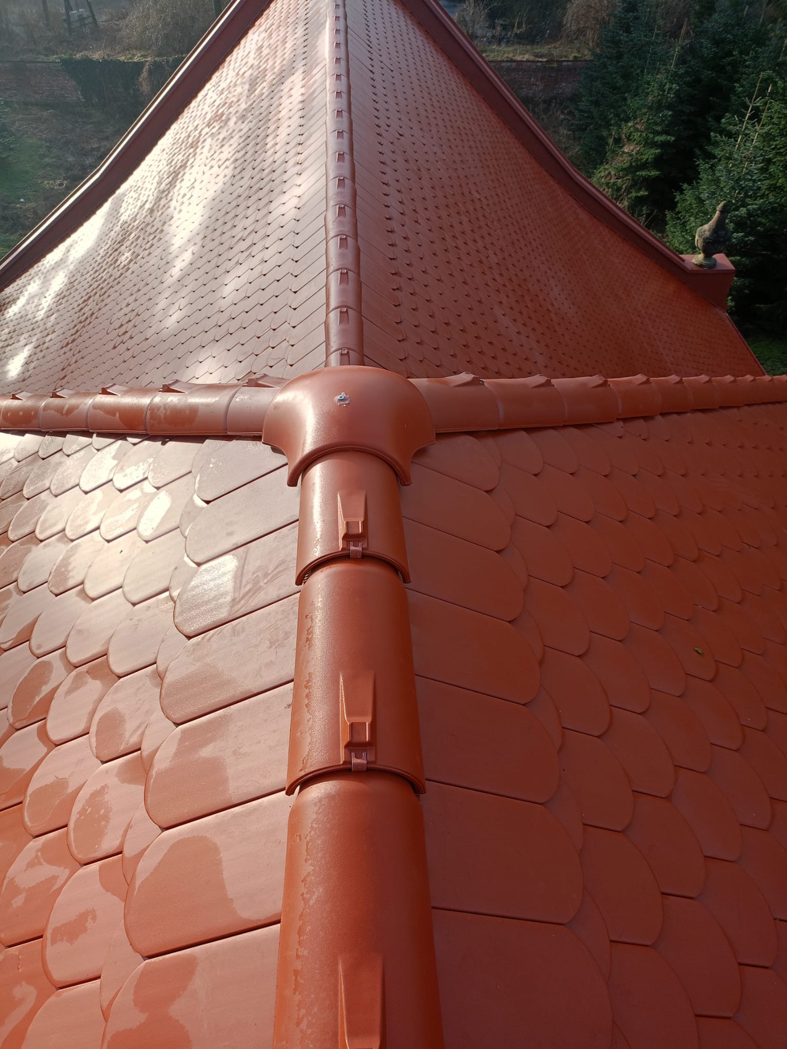 Czerwony dach z ceramicznej dachówki karpiówki z systemem rynnowym w kolorze, widok z góry na połączenie rynien, w tle zieleń drzew i krzewów.