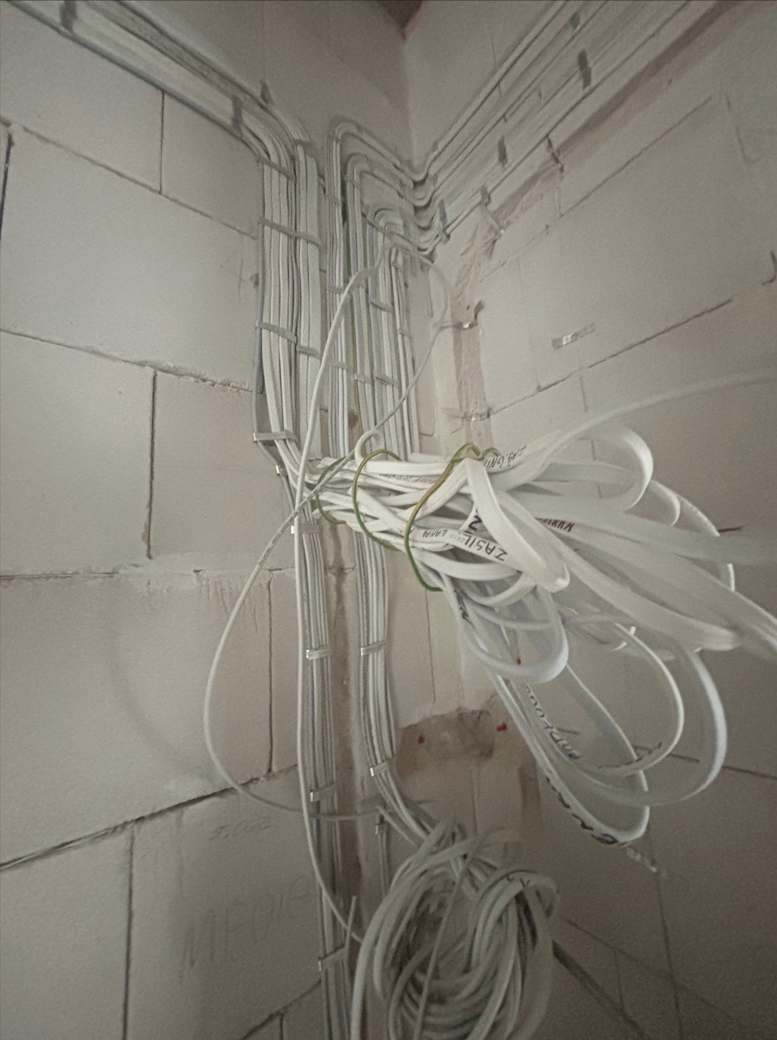 Wiązka kabli elektrycznych prowadzona w korytkach na surowej ścianie z bloczków, tymczasowo związana zielonym sznurkiem, widoczne napisy na kablach.