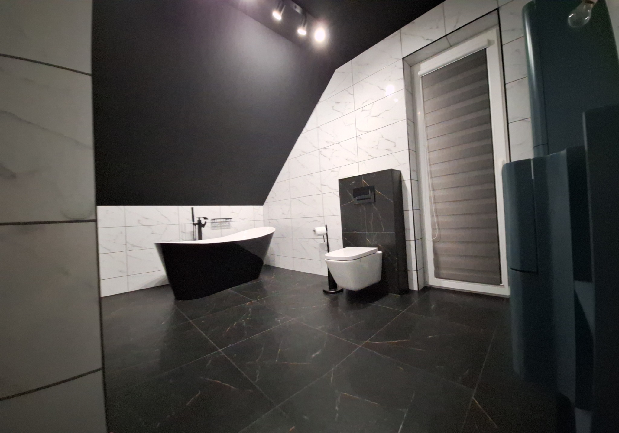 Nowoczesna łazienka z czarno-białymi płytkami, wolnostojącą wanną i czarną armaturą. Uwagę przykuwa czarna, skośna ściana pod sufitem i marmurowa podłoga.