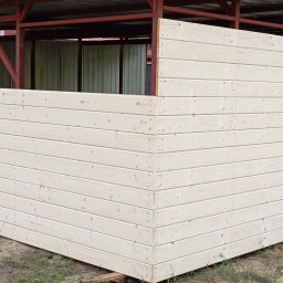 Kamil Woźniak - Fragment drewnianej konstrukcji z metalowym zadaszeniem, widoczne poziome deski i metalowe słupy, w tle zieleń