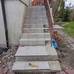 Kamil Woźniak - Układanie szarych płytek na schodach zewnętrznych, widoczny poziomica, szczotka do fugowania i kabel zasilający