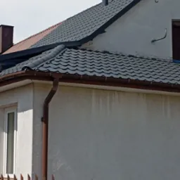 Fragment elewacji domu z szarą dach&oacute;wką, brązowymi rynnami i metalowym kominem, widoczne okno i fragment ogrodzenia.