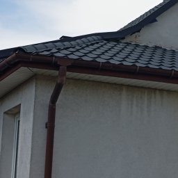 Kamil Woźniak - Fragment elewacji budynku z szarym tynkiem, ciemnoszarą dachówką i brązową rynną spustową. Widoczne zabrudzenia na ścianie pod rynną.