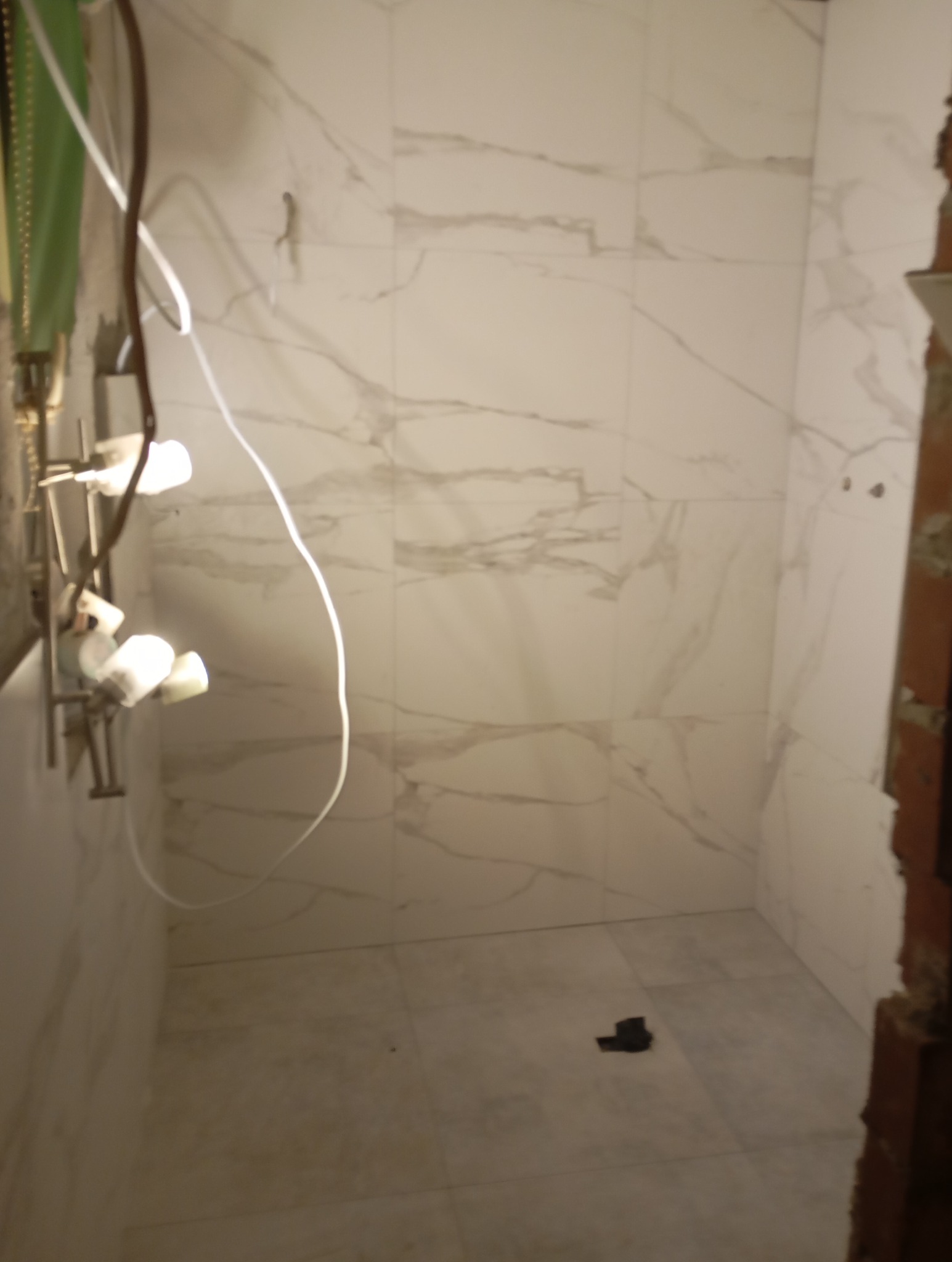 Wnętrze łazienki w trakcie prac wykończeniowych, widoczne płytki imitujące marmur na ścianach i podłodze, niedokończone obramowanie drzwi i wystające kable.