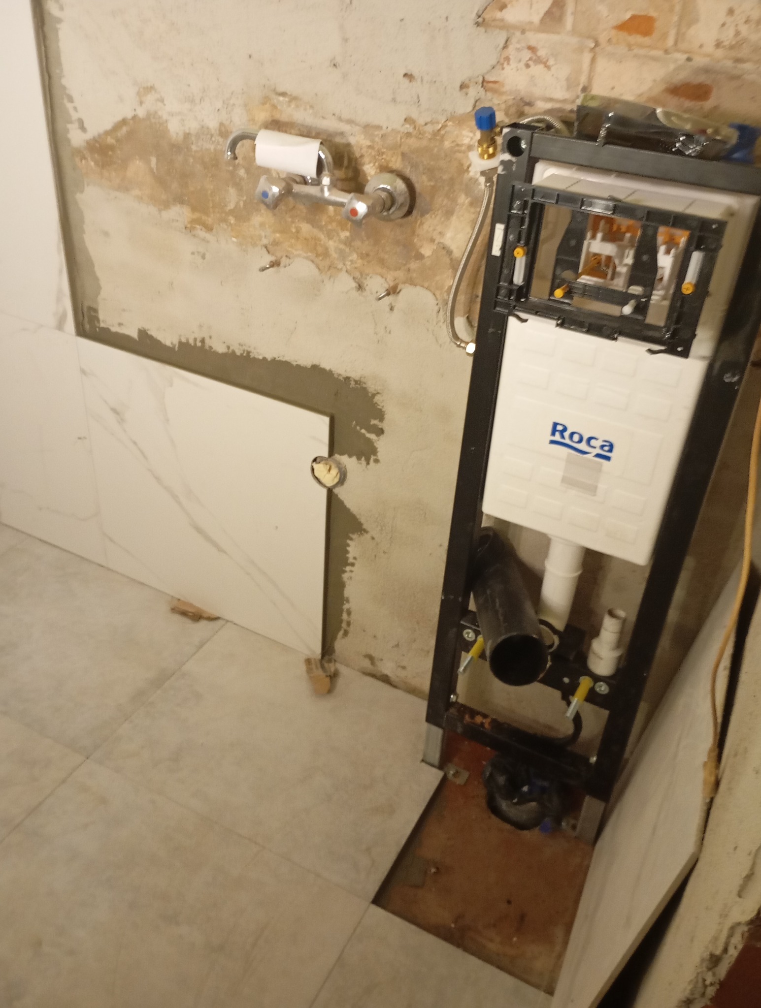 Wnęka w trakcie wykańczania z zamontowanym stelażem podtynkowym WC marki Roca, widoczne fragmenty płytek na ścianie i podłodze oraz surowa ściana z instalacją wodną.