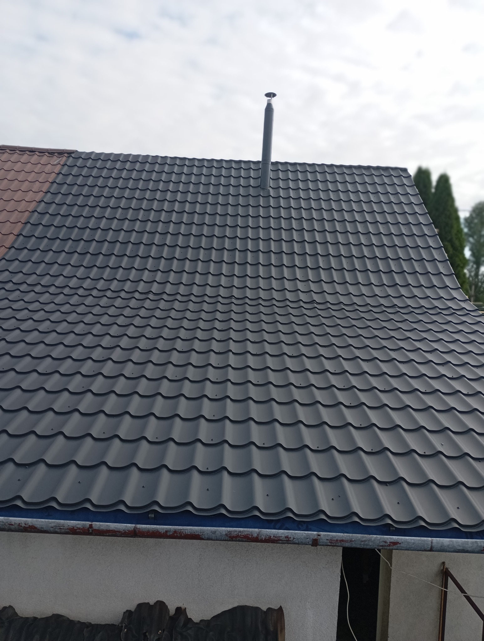 Nowe, grafitowe pokrycie dachowe z blachy trapezowej z kominem wentylacyjnym, widok z dołu na tle pochmurnego nieba.