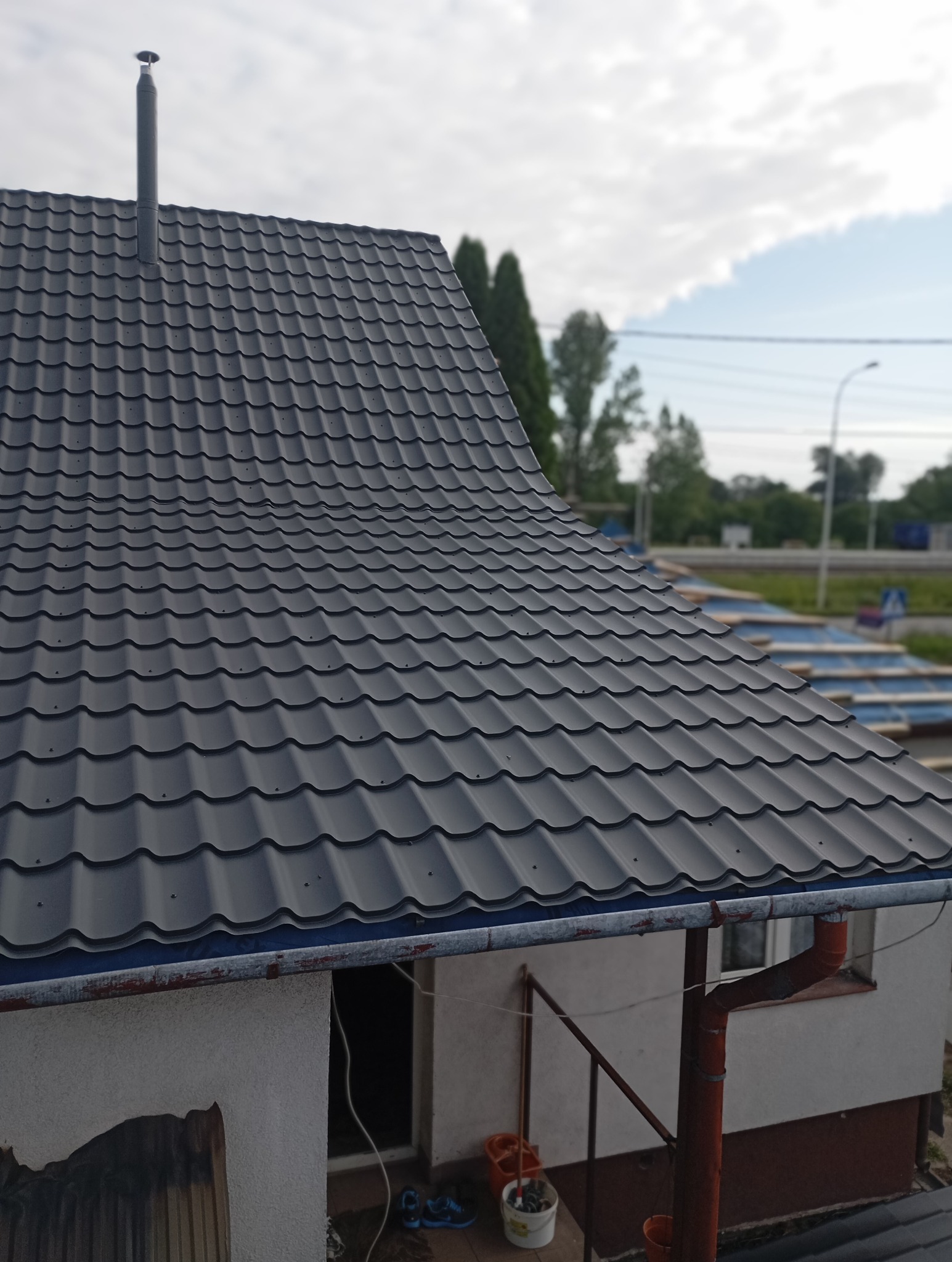 Nowy, ciemnoszary dach z blachy trapezowej z kominem, widoczny z góry, z elementami orynnowania i fragmentem ściany budynku w tle.