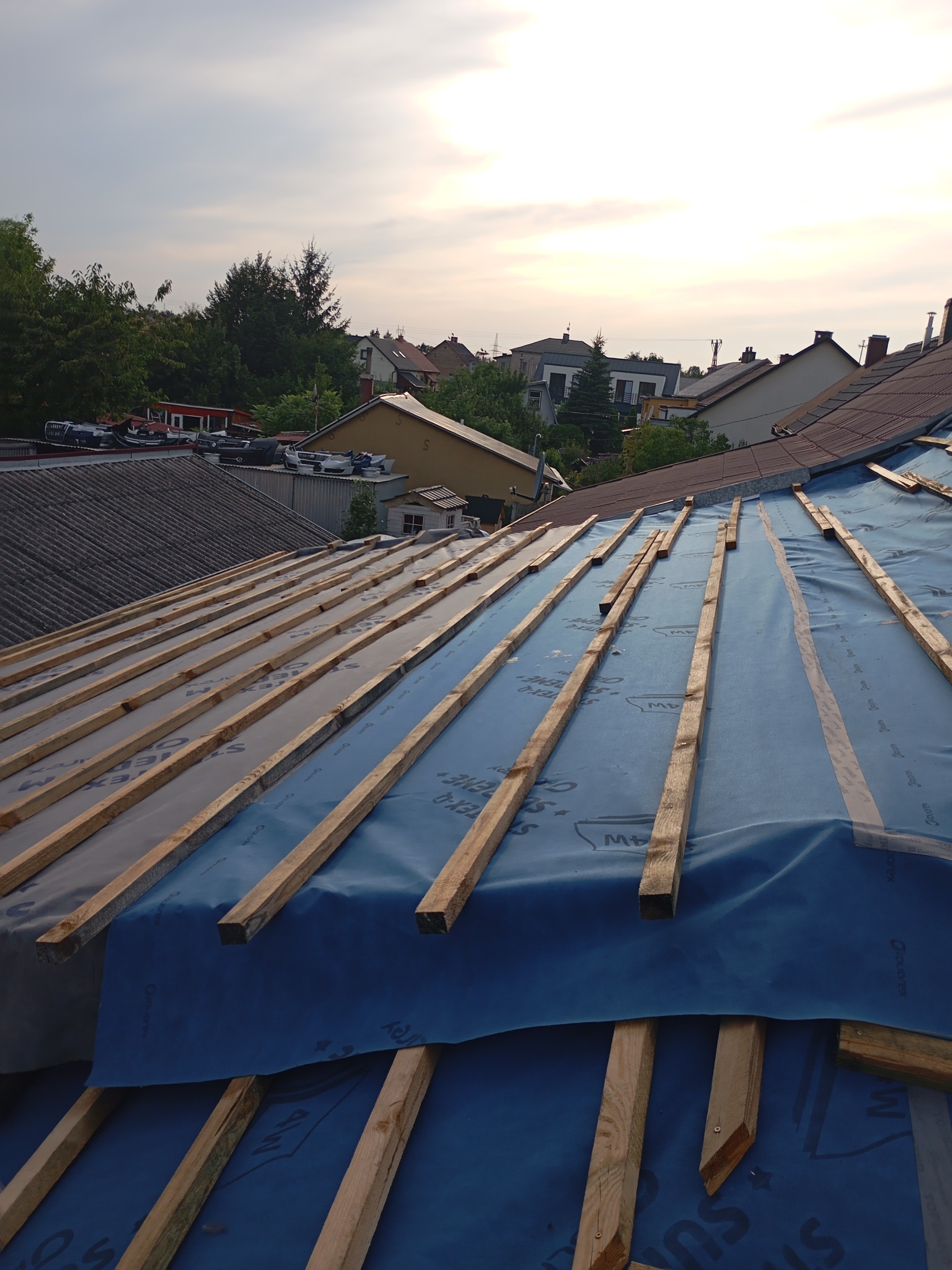 Widok z góry na konstrukcję dachu pokrytą niebieską membraną i drewnianymi łatami, przygotowaną pod dalsze prace dekarskie, z widocznymi domami i drzewami w tle.