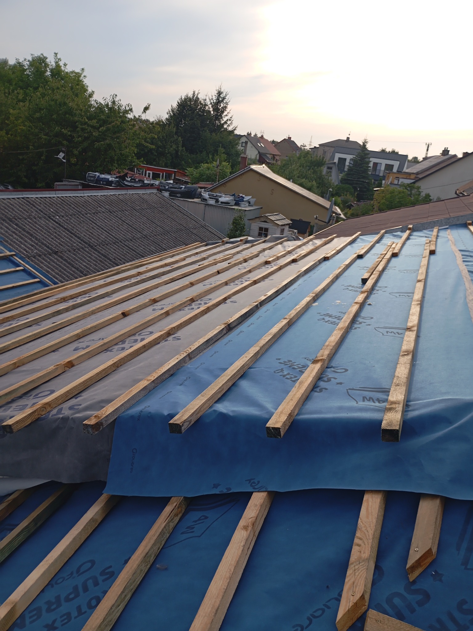 Konstrukcja dachu w trakcie budowy z widoczną niebieską membraną i drewnianymi łatami, na tle zabudowań mieszkalnych i drzew.