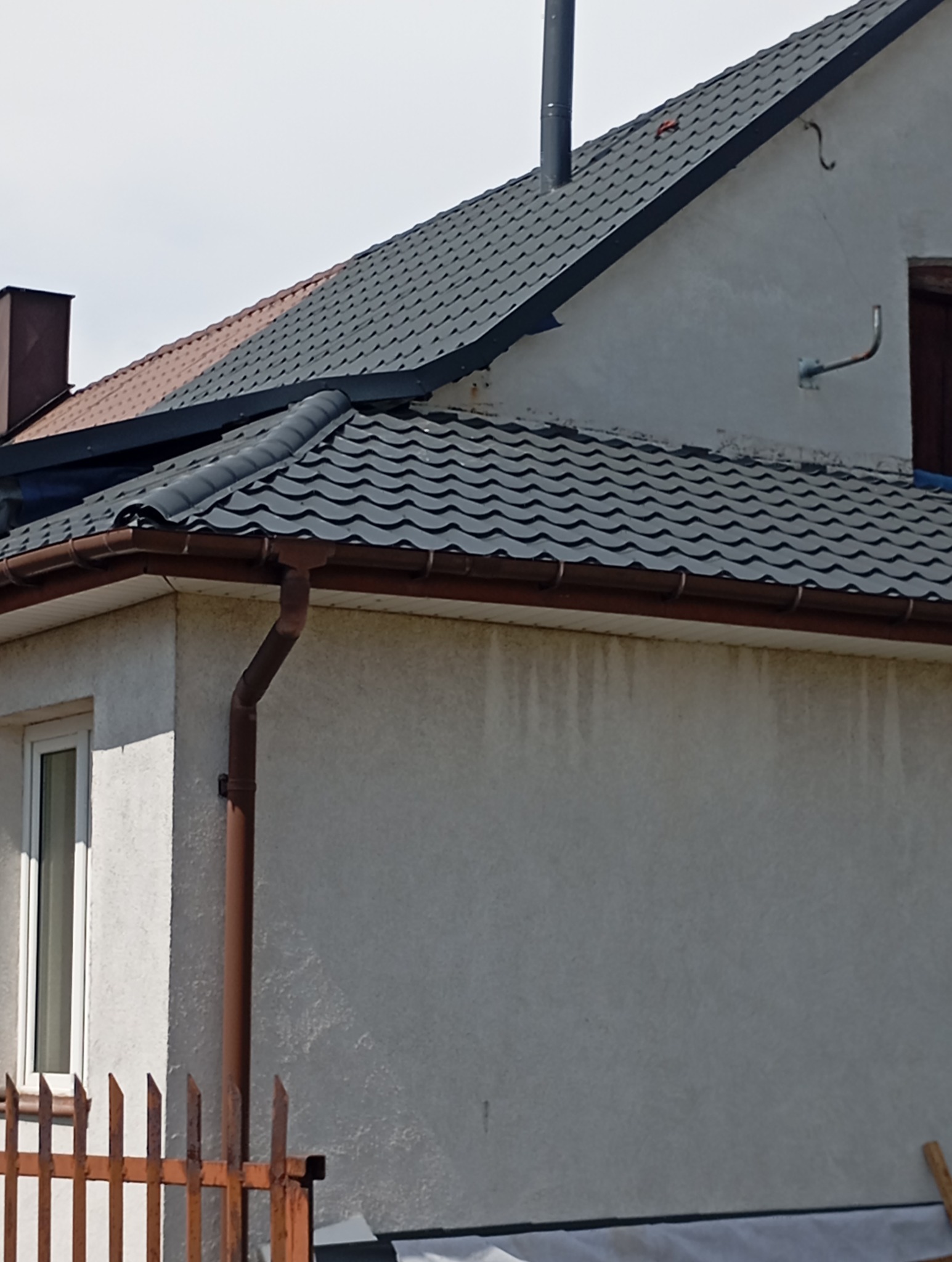 Fragment elewacji domu z szarą dachówką, brązowymi rynnami i metalowym kominem, widoczne okno i fragment ogrodzenia.