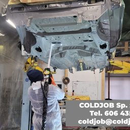 COLDJOB SPÓŁKA Z OGRANICZONĄ ODPOWIEDZIALNOŚCIĄ - Pracownik w kombinezonie ochronnym, z maską i rękawicami, czyści podwozie samochodu podniesionego na podnośniku w warsztacie.