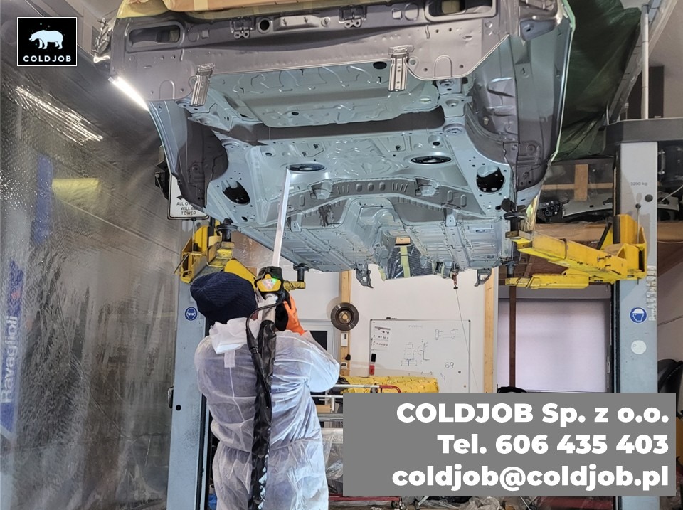 Pracownik w kombinezonie ochronnym, z maską i rękawicami, czyści podwozie samochodu podniesionego na podnośniku w warsztacie.