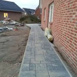 Bud Max - Świeżo ułożony chodnik z szarej kostki brukowej o zróżnicowanych kształtach, biegnący wzdłuż ceglanej ściany budynku. W tle widoczny plac budowy z materiałami i kolejny budynek w trakcie budowy.