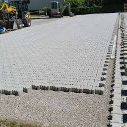 Bud Max - Jasnoszara kostka brukowa ułożona na dużej powierzchni, z widocznymi maszynami budowlanymi w tle i rzędem kostek przygotowanych do ułożenia na krawędzi.