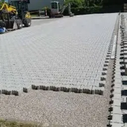 Jasnoszara kostka brukowa ułożona na dużej powierzchni, z widocznymi maszynami budowlanymi w tle i rzędem kostek przygotowanych do ułożenia na krawędzi.