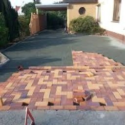 Bud Max - Częściowo ułożona, prostokątna kostka brukowa w odcieniach czerwieni i beżu na podjeździe do domu. Widoczne przygotowane podłoże z czarnej geowłókniny. Fragmenty kostki nieułożone, leżące obok.