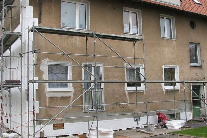 Ocieplanie budynku styropianem w Bielawie, widoczne rusztowania i pracownik w czerwonym kombinezonie podczas montażu izolacji.