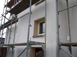 Ocieplony budynek w trakcie prac wykończeniowych, widoczne rusztowania i przygotowane okno do dalszych prac.